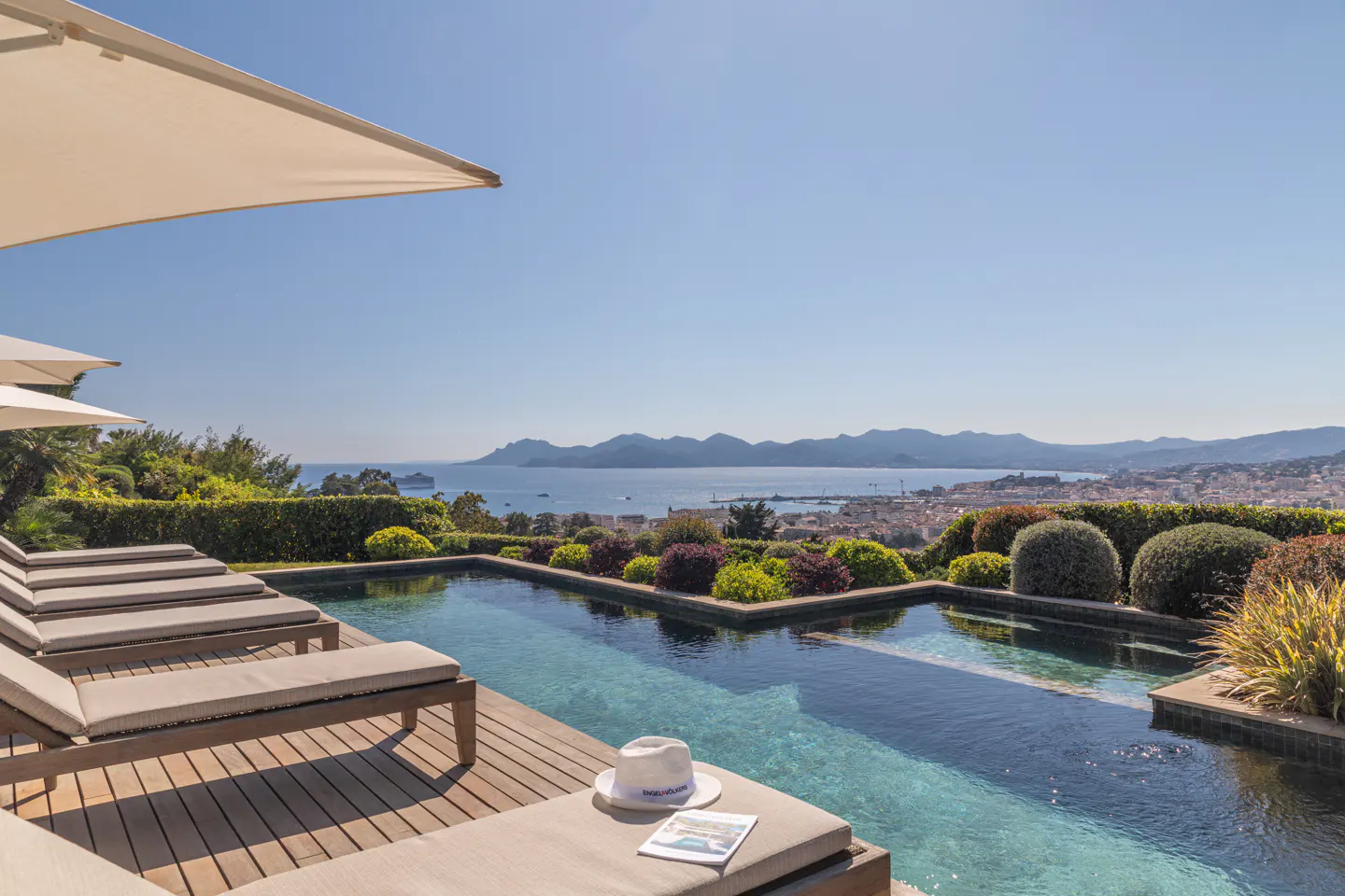 Luxury villa with pool overlooking the sea. Lounge chairs and umbrellas on the wooden deck. Hat and magazine on the chair.