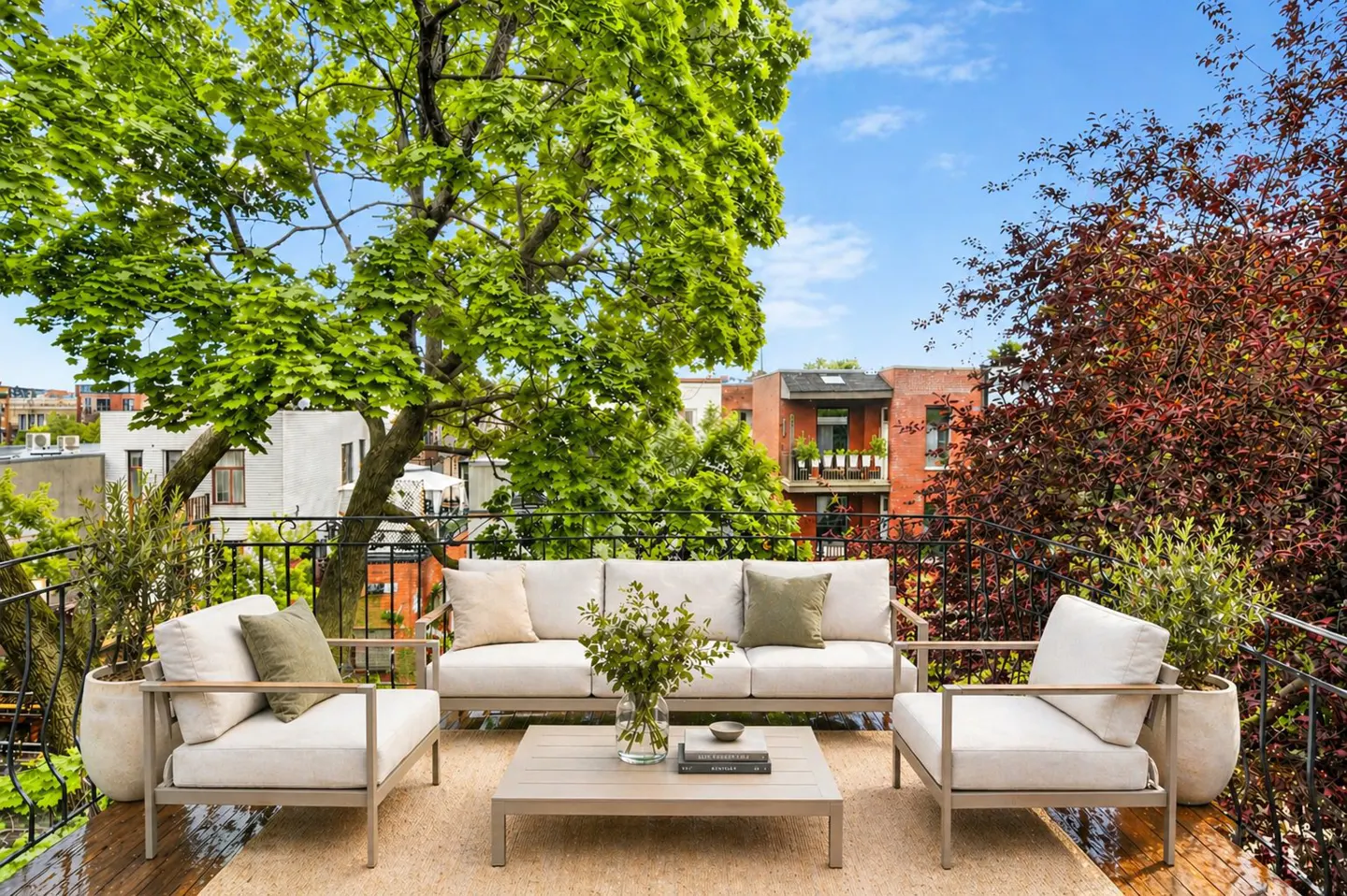 Outdoor patio with a beige sofa, two chairs, and a coffee table on a rug. Trees and buildings are in the background.