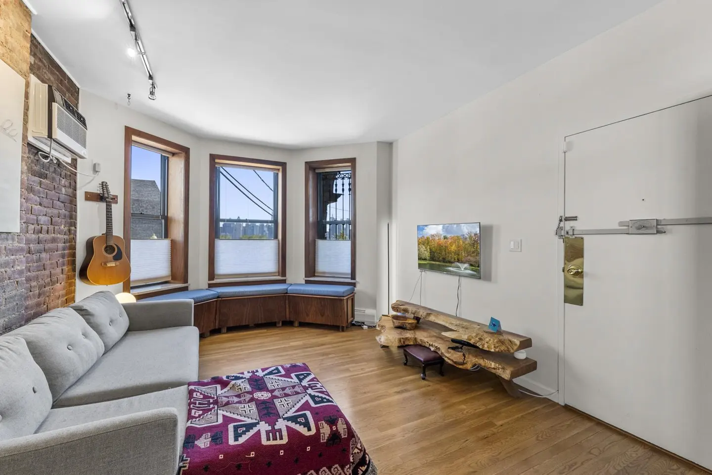 Bright living room with a gray sofa, brick wall, and bay window with built-in seating. A guitar hangs on the wall, and a unique wood table sits beneath a TV.
