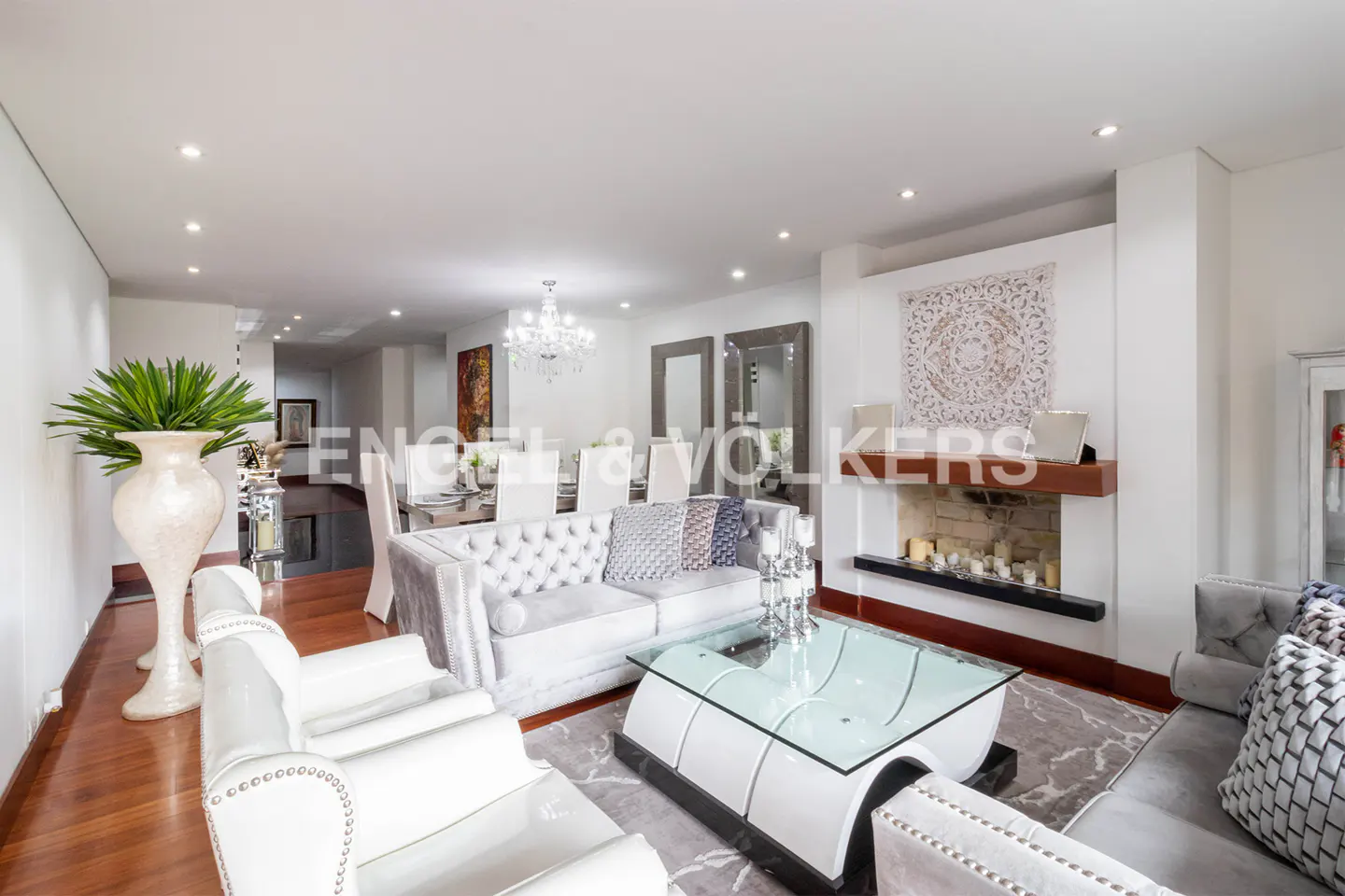 Bright living room with white walls, hardwood floors, and gray sofas. A glass coffee table sits on a gray rug. A fireplace and dining area are in the background.