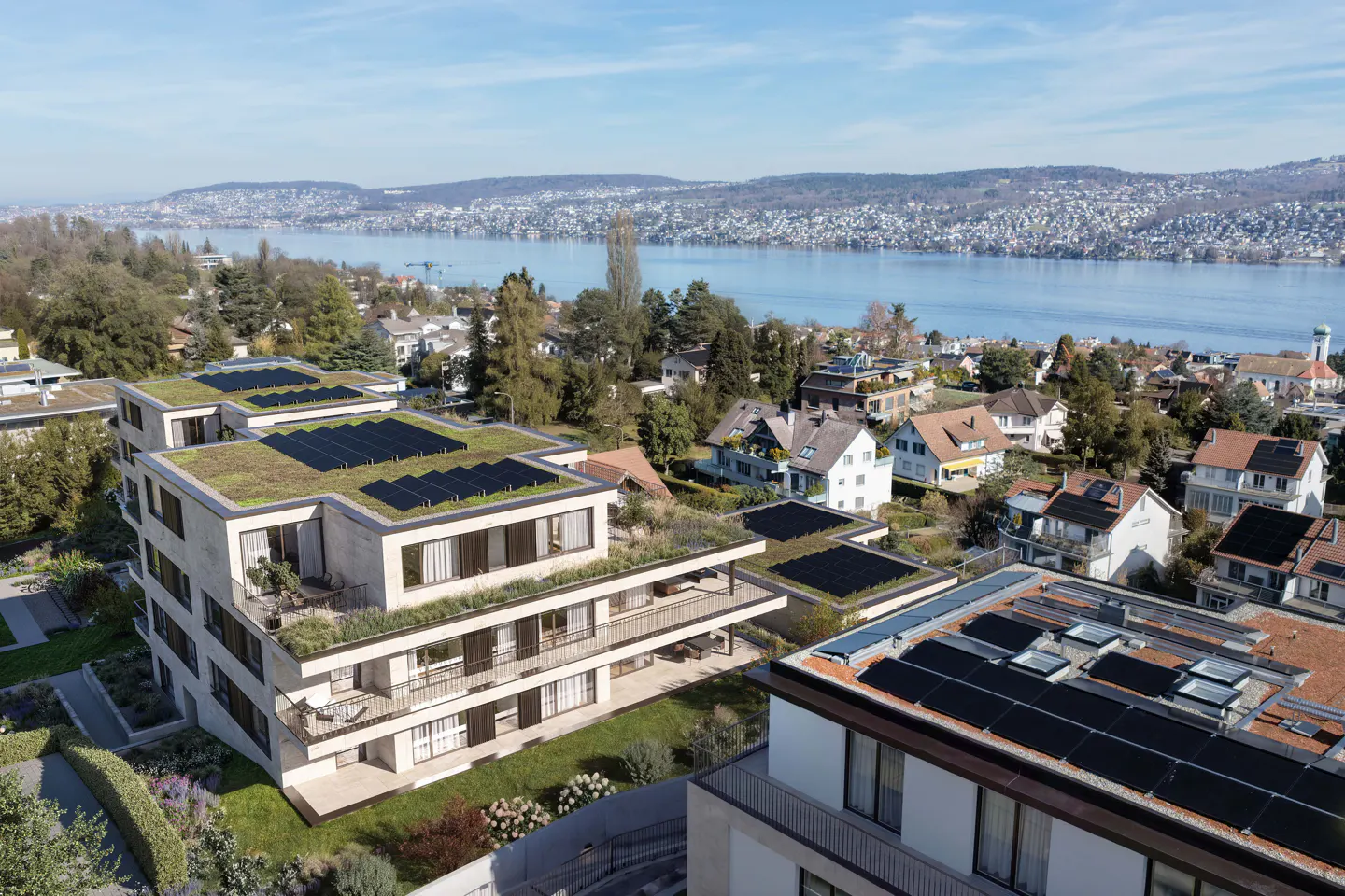 Aerial view of modern apartments with green roofs and solar panels, overlooking a lake and distant hills.