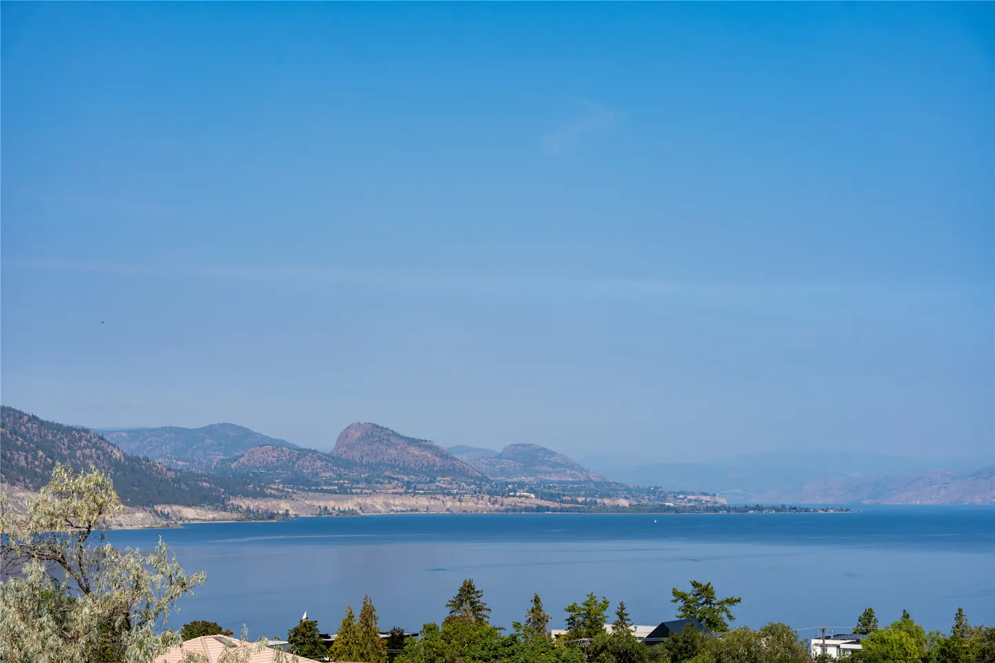 Scenic view of a blue lake with mountains in the background under a clear blue sky. Green trees are visible in the foreground.
