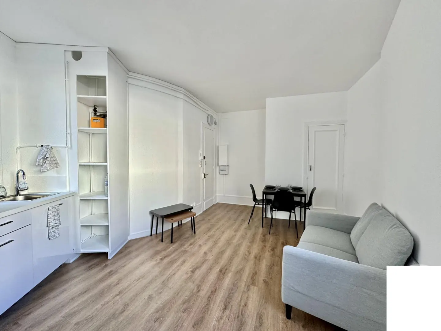 Bright studio apartment with wood floors, white walls, a gray sofa, and a black dining table with four chairs.