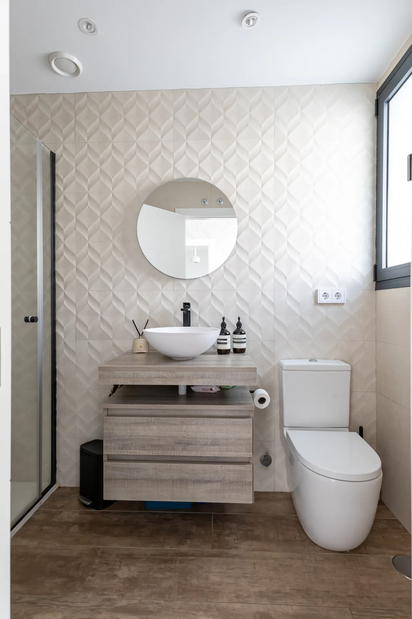 Bathroom with a white toilet, sink, oval mirror, and textured beige wall tiles. Wood-look flooring and a shower are also visible.