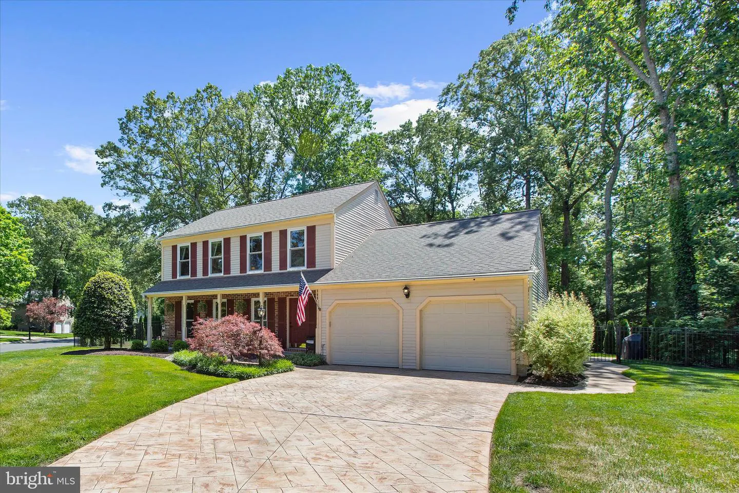 Two-story beige house with red shutters, a two-car garage, and a patterned driveway on a sunny day.
