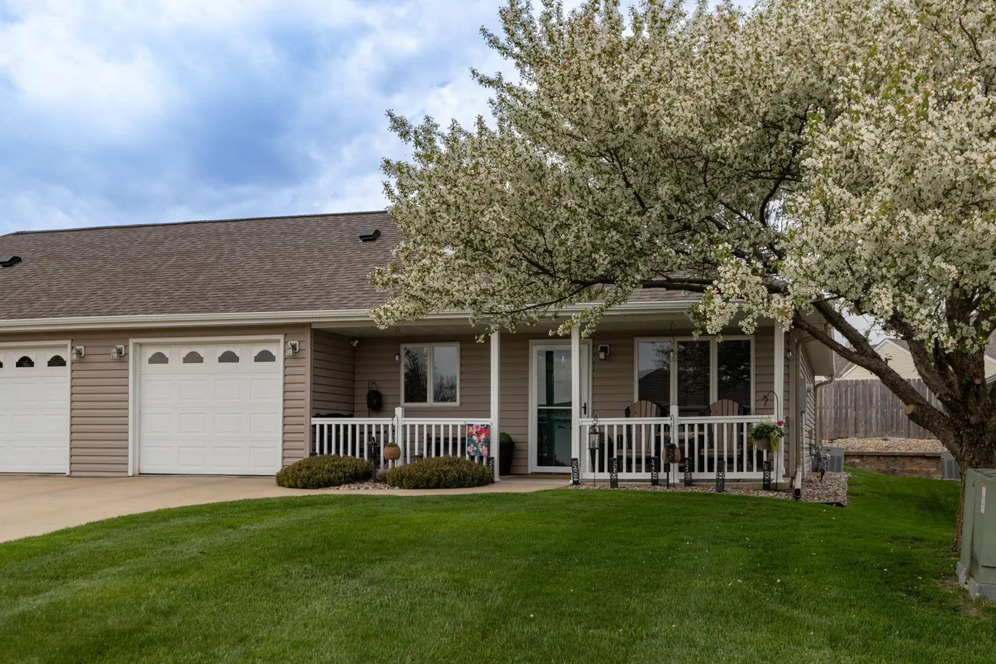 Tan house with white garage doors and a porch with white railings. A flowering tree is in the yard.