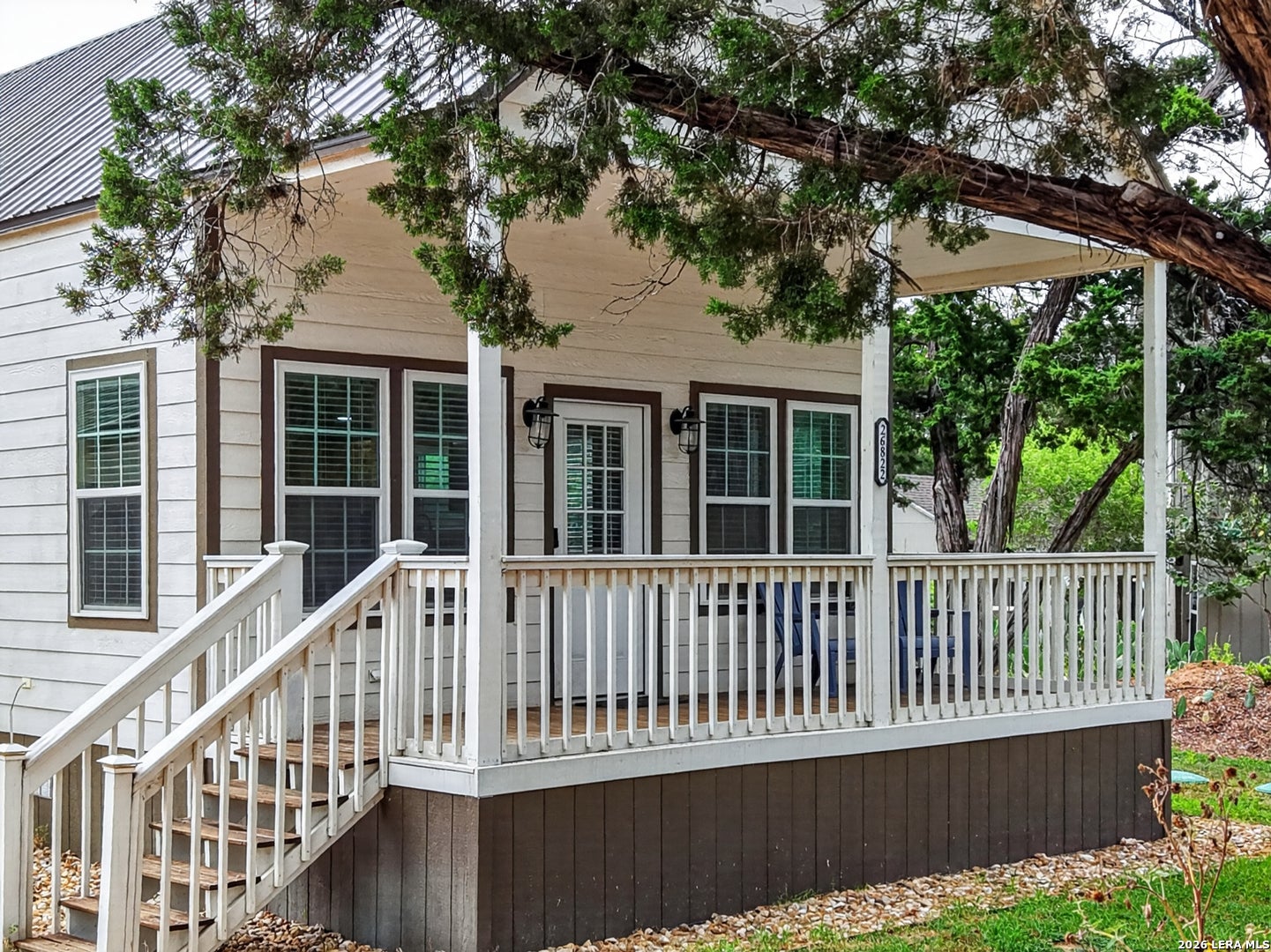 White cottage with a covered front porch, wooden stairs, dark trim, and a metal roof, framed by a large tree branch. [AI-generated text]