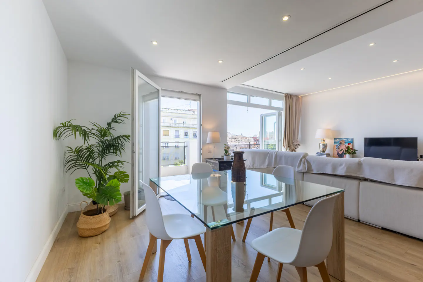 Bright, modern apartment interior with a glass table, white chairs, and open balcony doors. A potted plant adds a touch of nature.