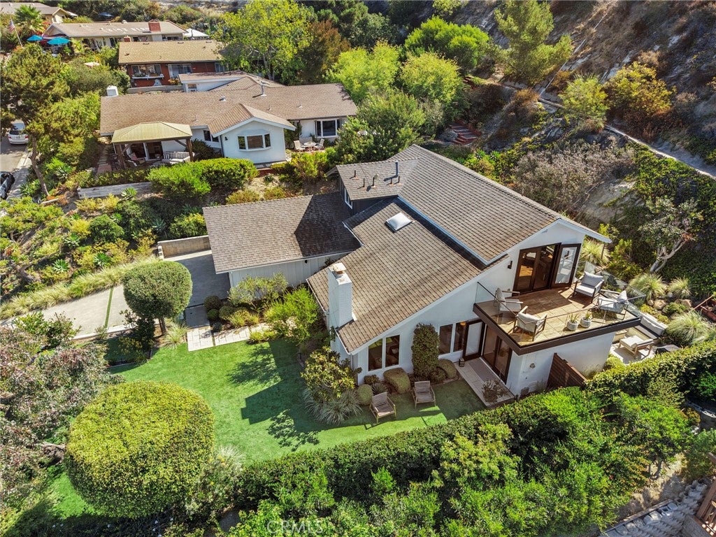 Aerial view of a white multi-level home with a brown roof, wooden deck, green lawn, and lush hillside landscaping. [AI-generated text]