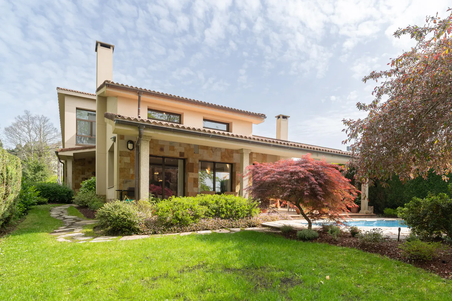 Two-story house with stone and stucco exterior, a red tile roof, and a green lawn. A swimming pool is visible in the background.