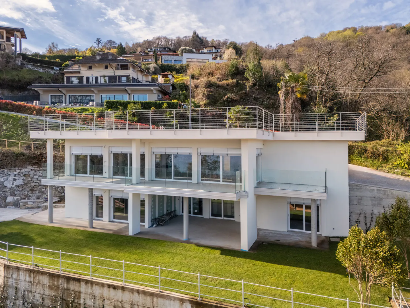 A modern, white two-story house with large windows and balconies sits on a green lawn, with hillside homes in the background.