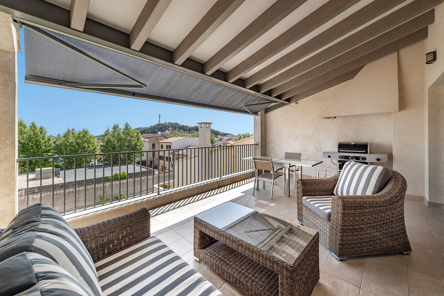 A covered patio with wicker furniture, a table, and a grill overlooks a town with trees and a hill in the distance.
