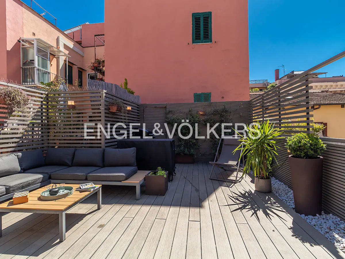 A rooftop terrace with gray outdoor furniture, potted plants, and a wooden deck. The building behind is pink with green shutters.