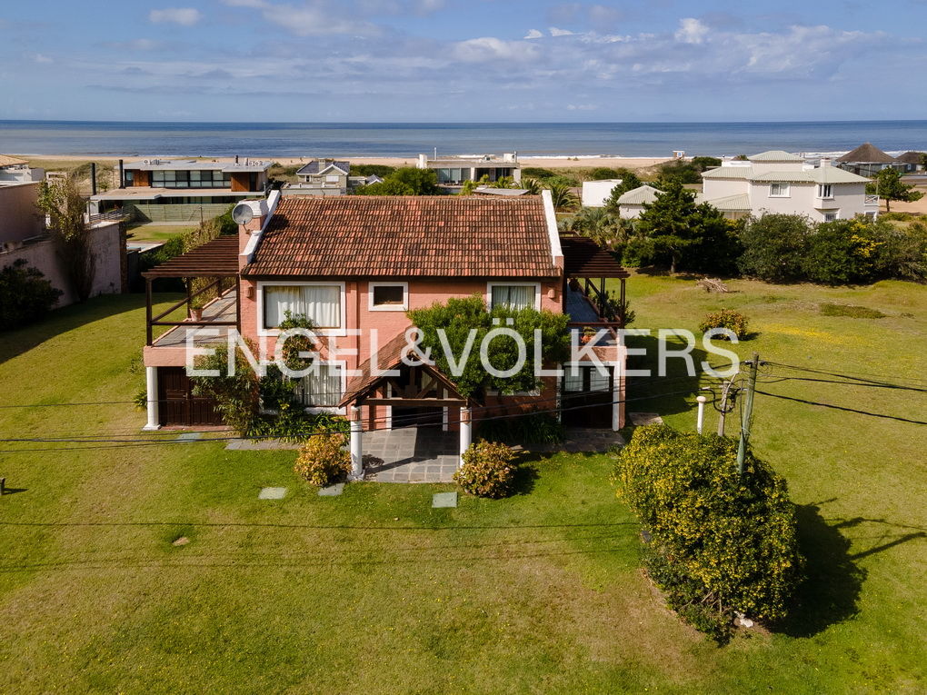Aerial view of a salmon-colored house with a red tile roof, green lawn, and ocean backdrop on a sunny day.