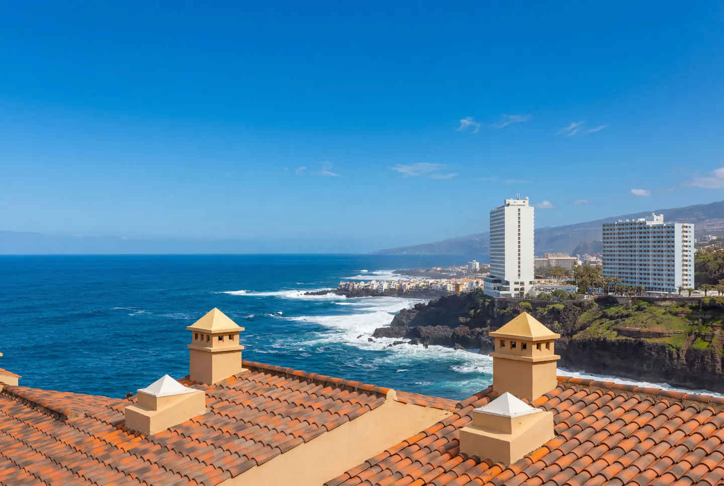 View from a terracotta tiled roof with chimneys, overlooking the ocean, coastline, and buildings under a clear blue sky.