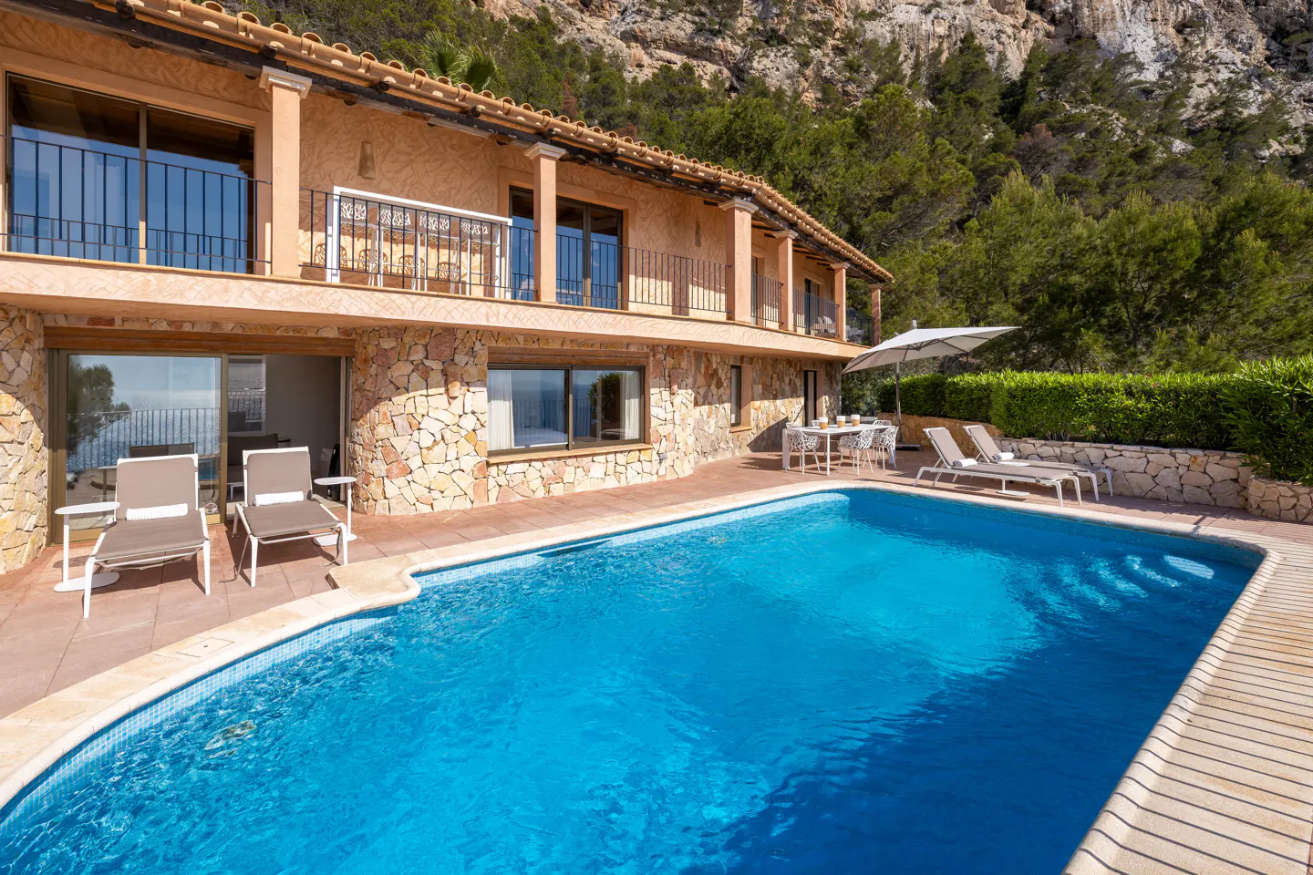 Two-story stone villa with a blue swimming pool, lounge chairs, and a patio dining area against a mountain backdrop.