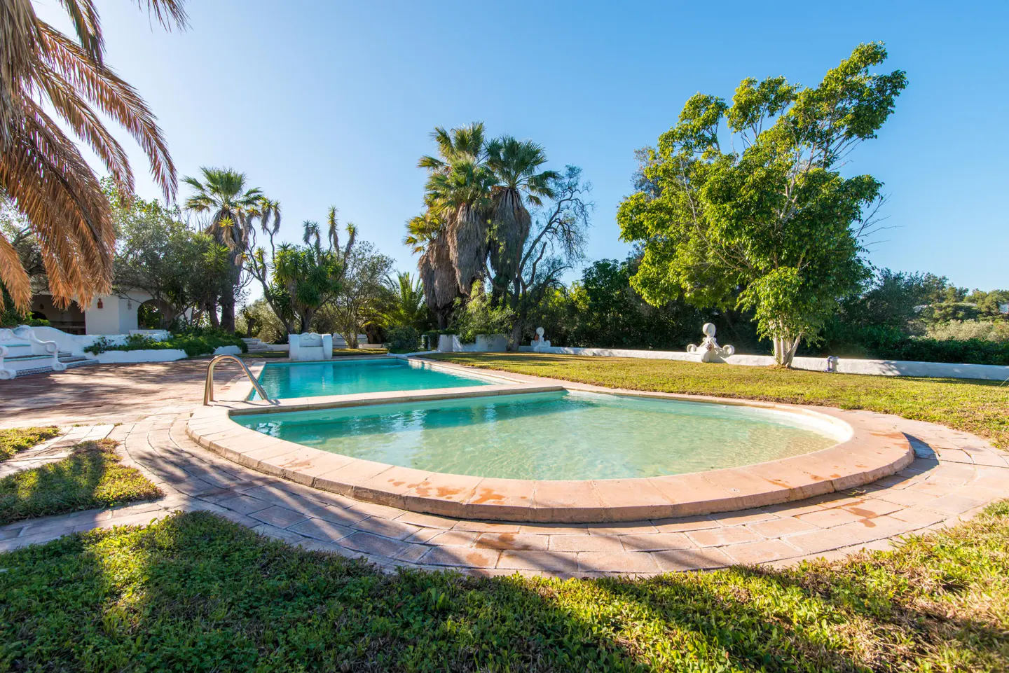 Outdoor pool with clear turquoise water, surrounded by stone and grass, with palm trees and blue sky.