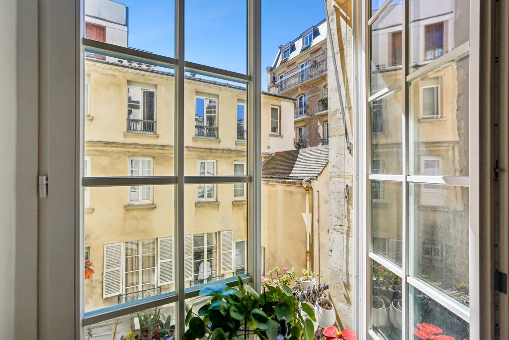View from an open window with white frames, showing buildings in a courtyard on a sunny day. Potted plants sit on the windowsill.