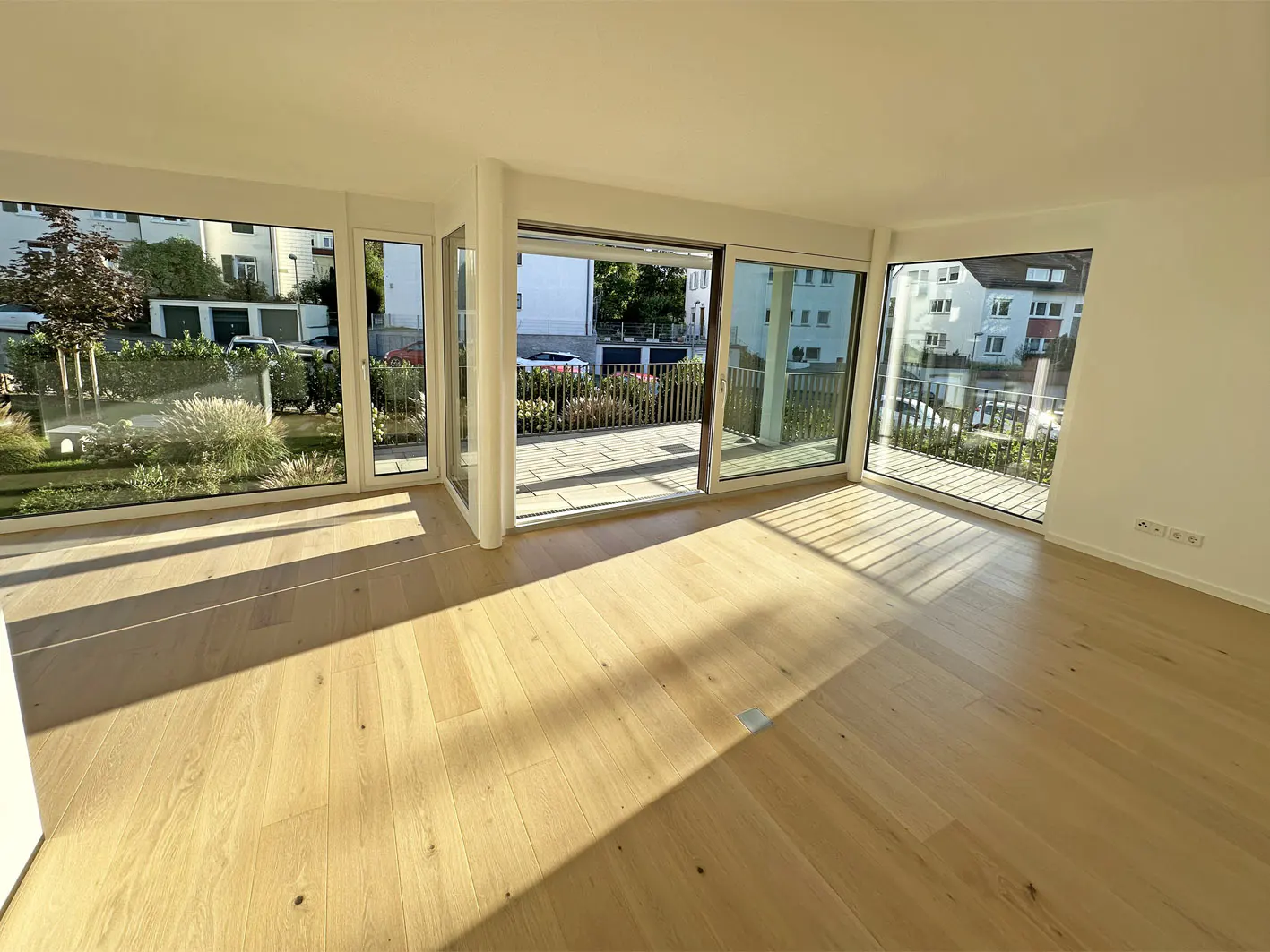 Bright, empty room with light wood floors and white walls. Large windows and sliding glass doors offer views of greenery and a patio.
