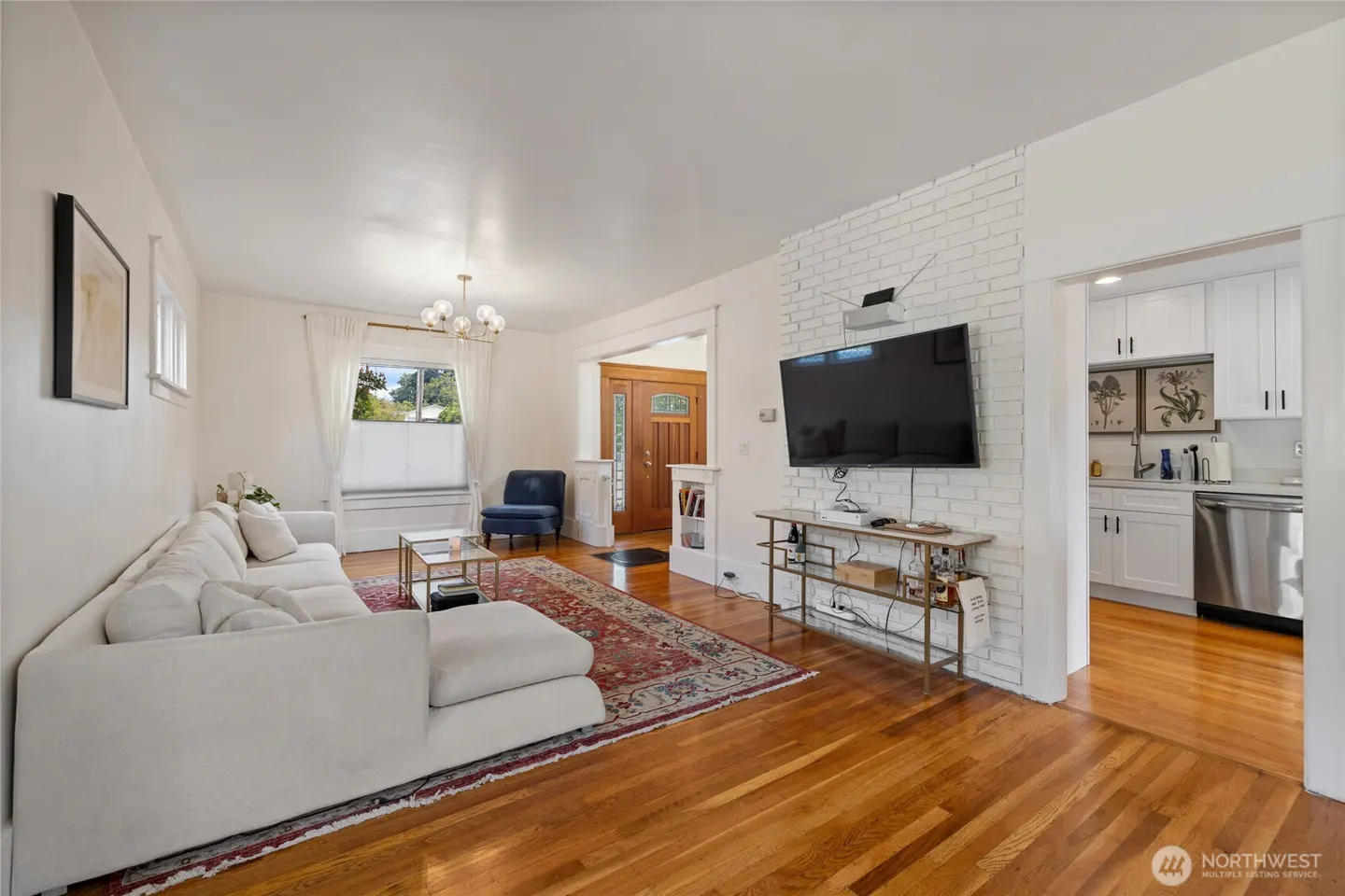 Bright living room with hardwood floors, white walls, and a large white sectional sofa. A TV is mounted on a white brick wall, and a kitchen is visible in the background.