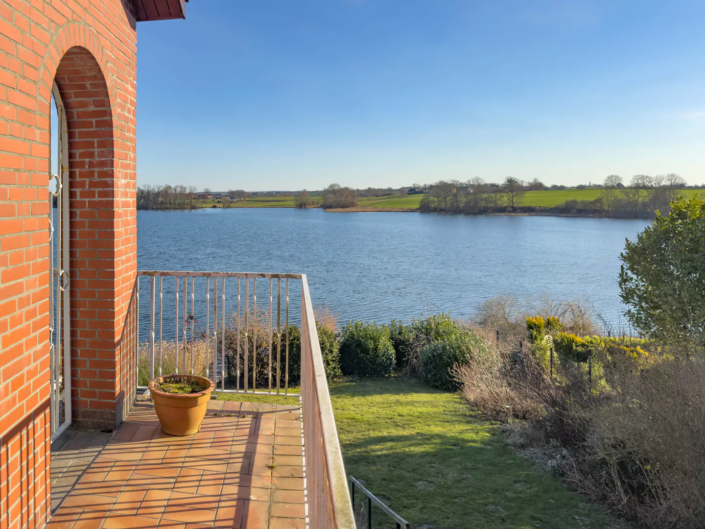 Balcony view of a lake from a brick house. A potted plant sits on the tiled balcony with a white metal railing. Green grass and trees surround the lake.