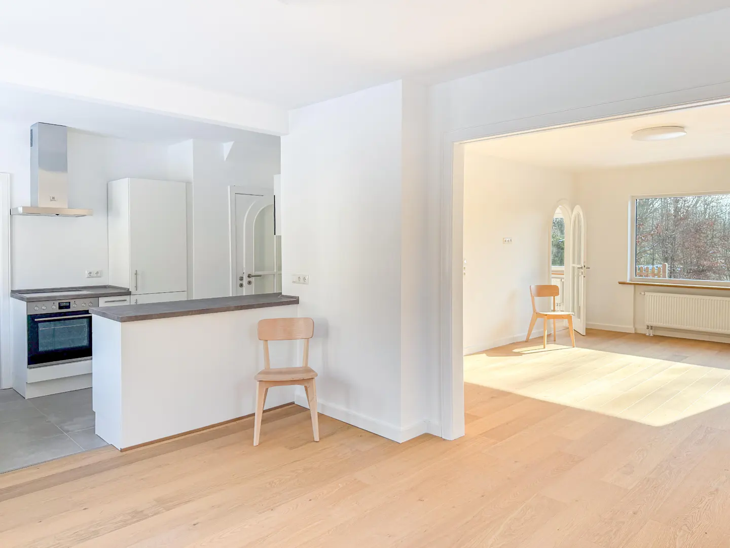 Bright, open-concept apartment with light wood floors. A white kitchen and a chair are visible. Another room is seen through a doorway.