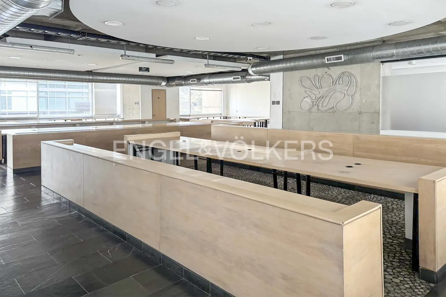 An empty office space with long wooden desks and black metal legs. The floor is dark gray tile, and there are large windows in the background.