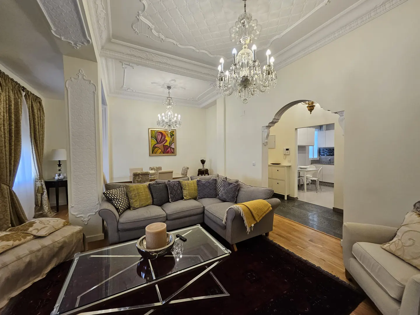 Elegant living room with a grey sectional sofa, glass coffee table, crystal chandeliers, and a view into the kitchen.