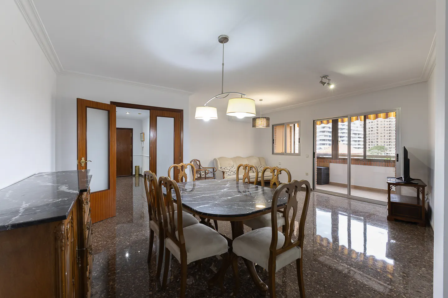 A dining room with a marble table, wooden chairs, and a view of a balcony with city buildings.