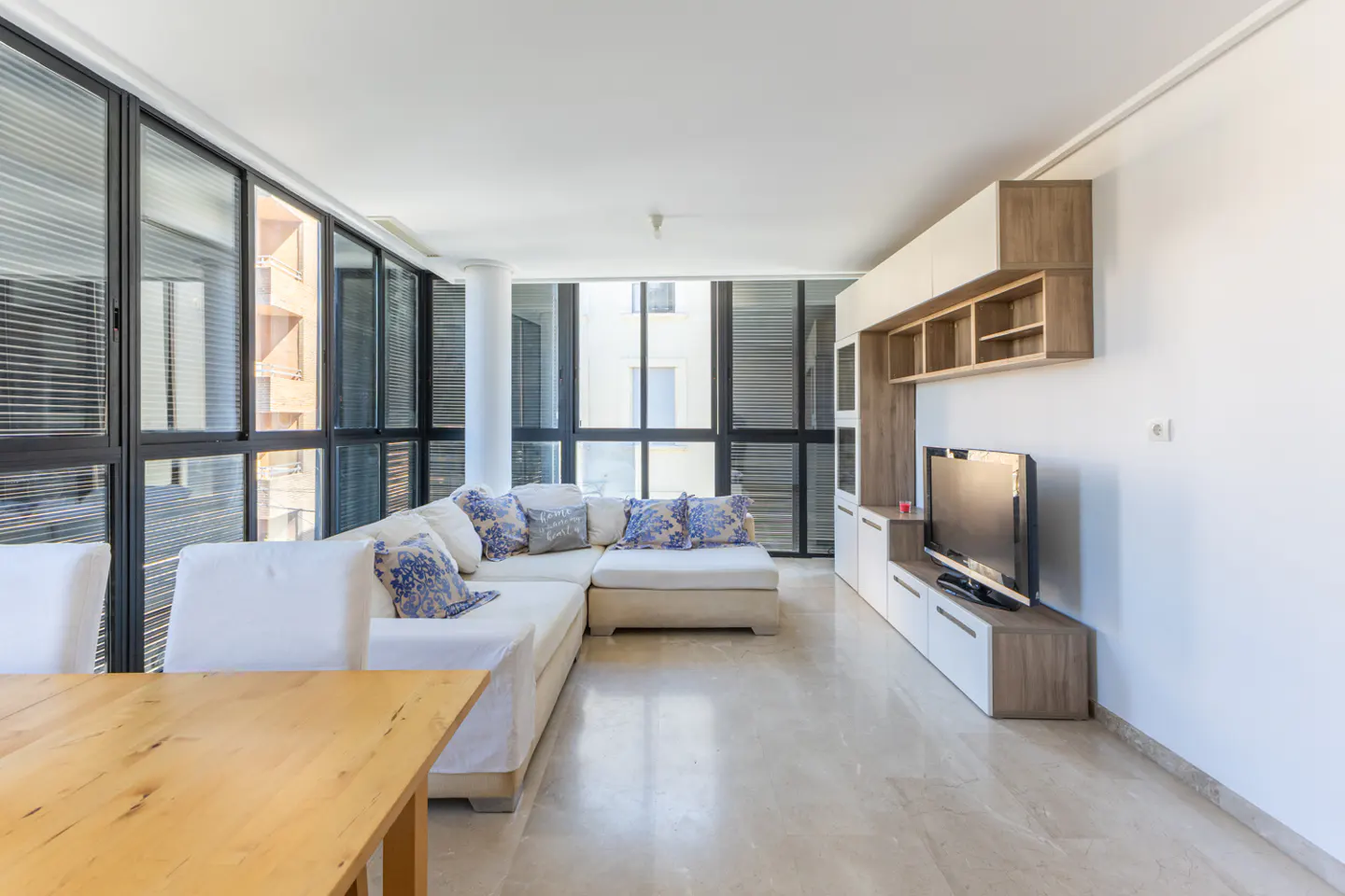 Bright living room with large windows, a white sectional sofa with blue pillows, and a wooden entertainment center with a TV.