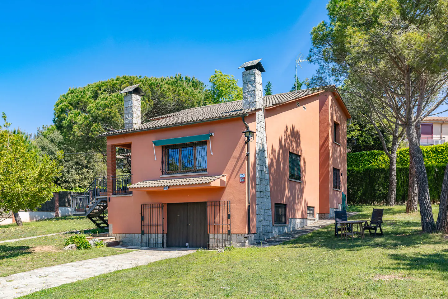 Two-story peach house with stone chimneys and a gated garage, surrounded by green lawn and trees under a blue sky.