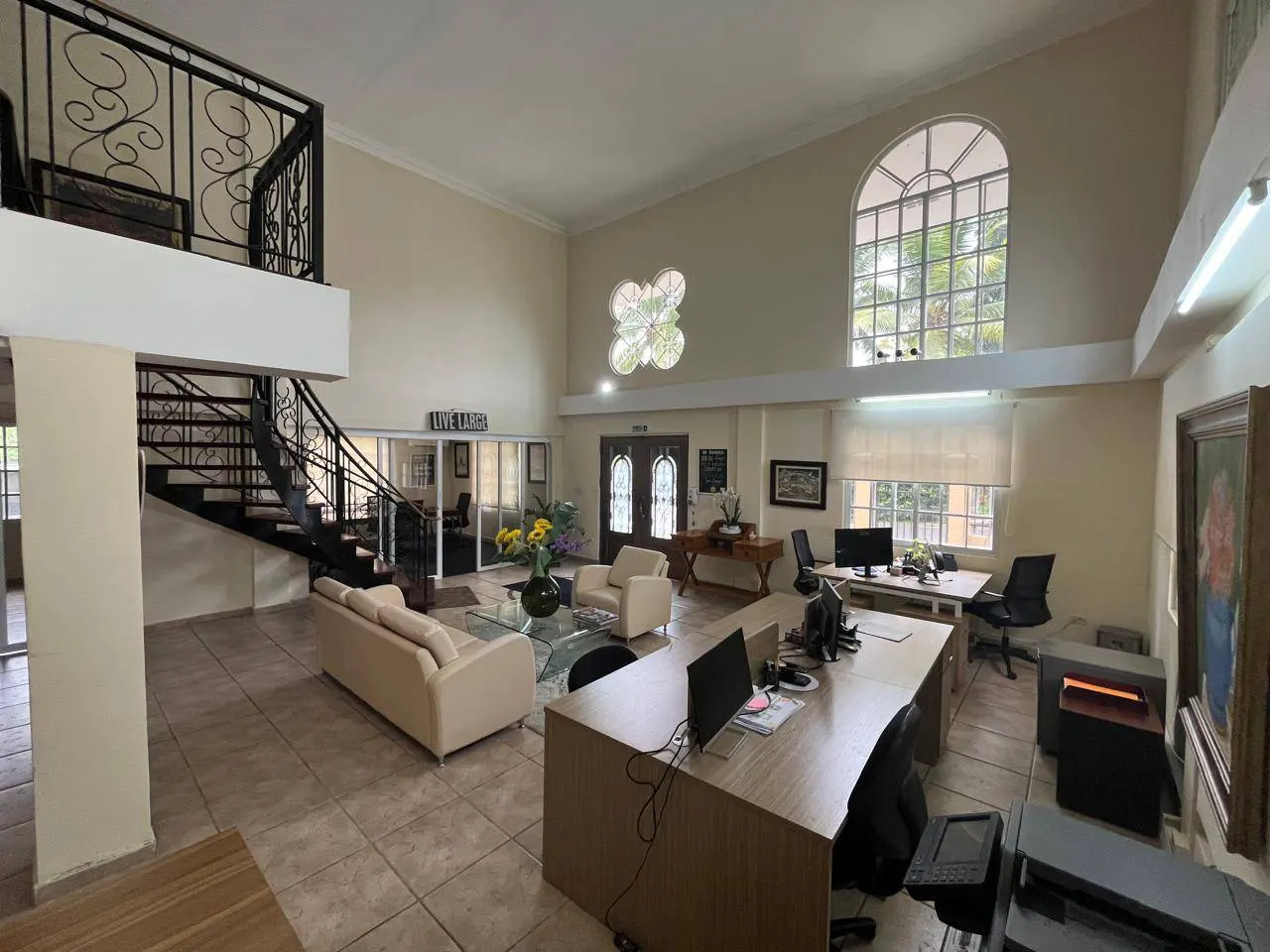 A real estate office with a staircase, desks, and a seating area. The walls are beige, and there is a large window.