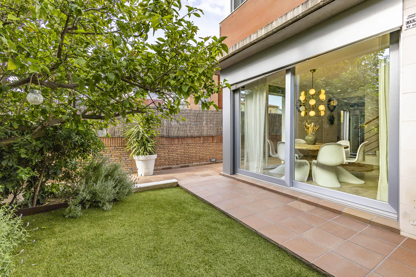 A patio with green grass, a tree, and a sliding glass door leading to a dining room with a round table and white chairs.