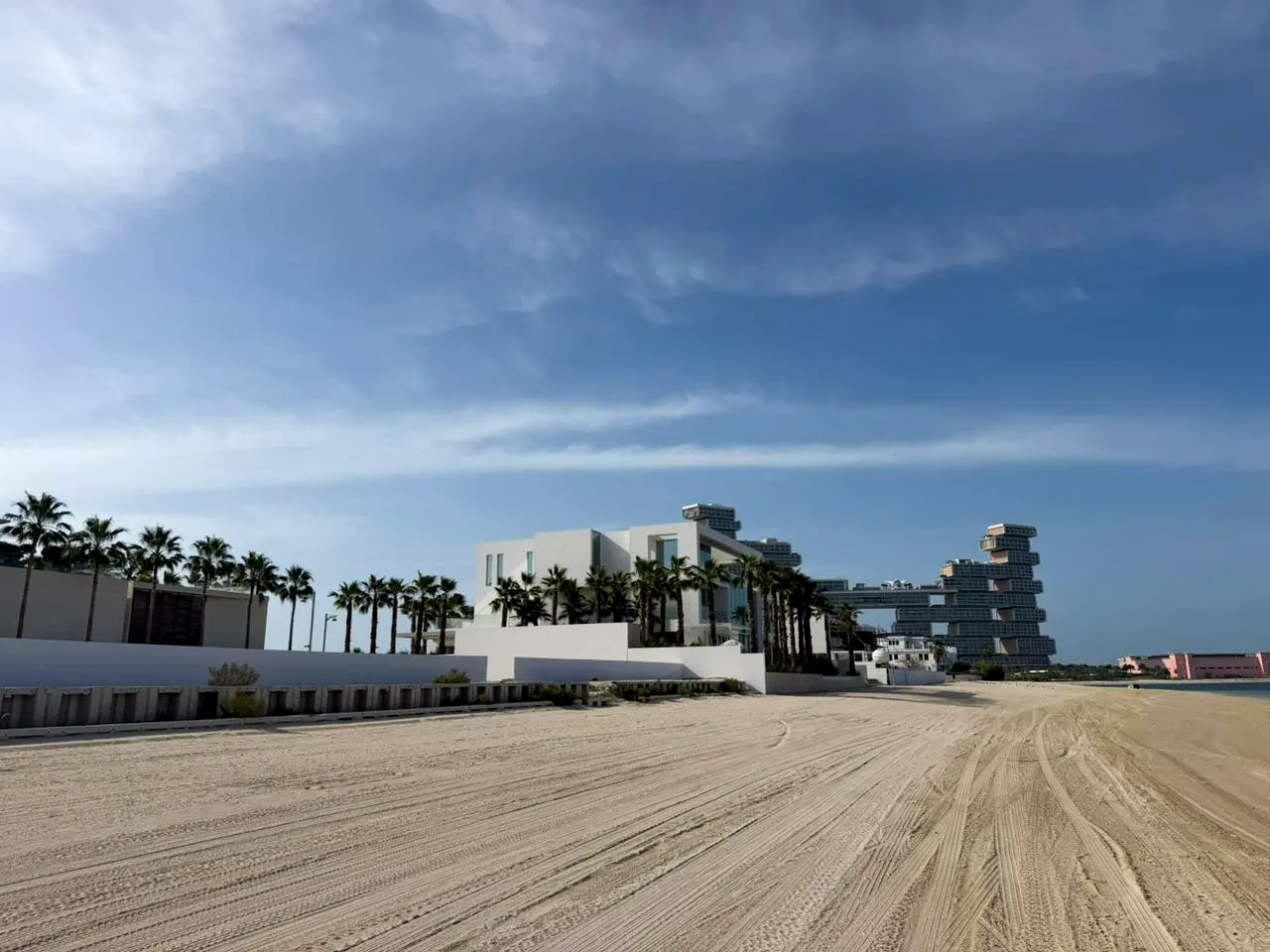 Beachfront property with modern white buildings, palm trees, and tire tracks in the sand under a blue sky.