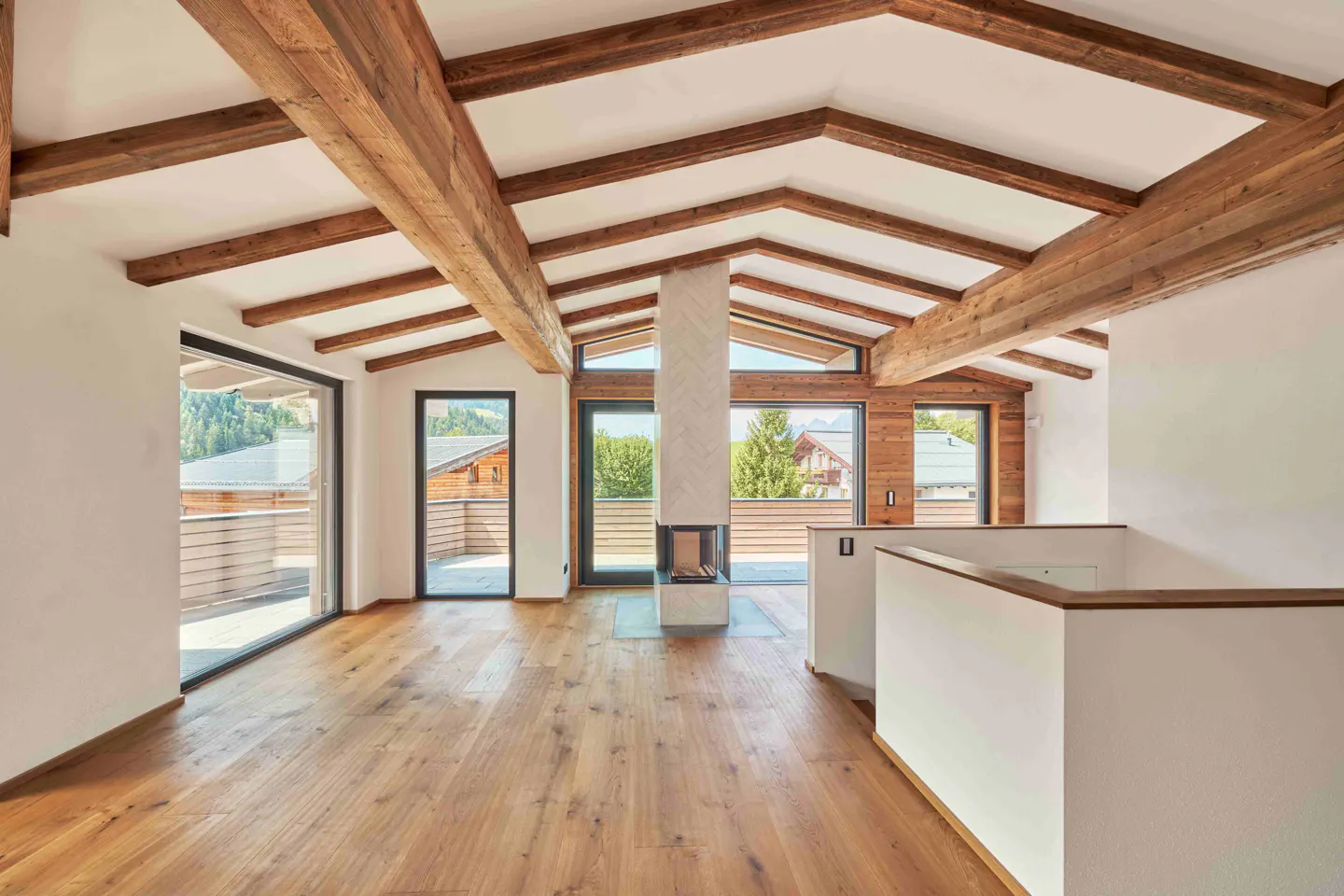 Bright, empty room with wood floors, exposed beams, and a modern fireplace. Large windows offer views of trees and houses.