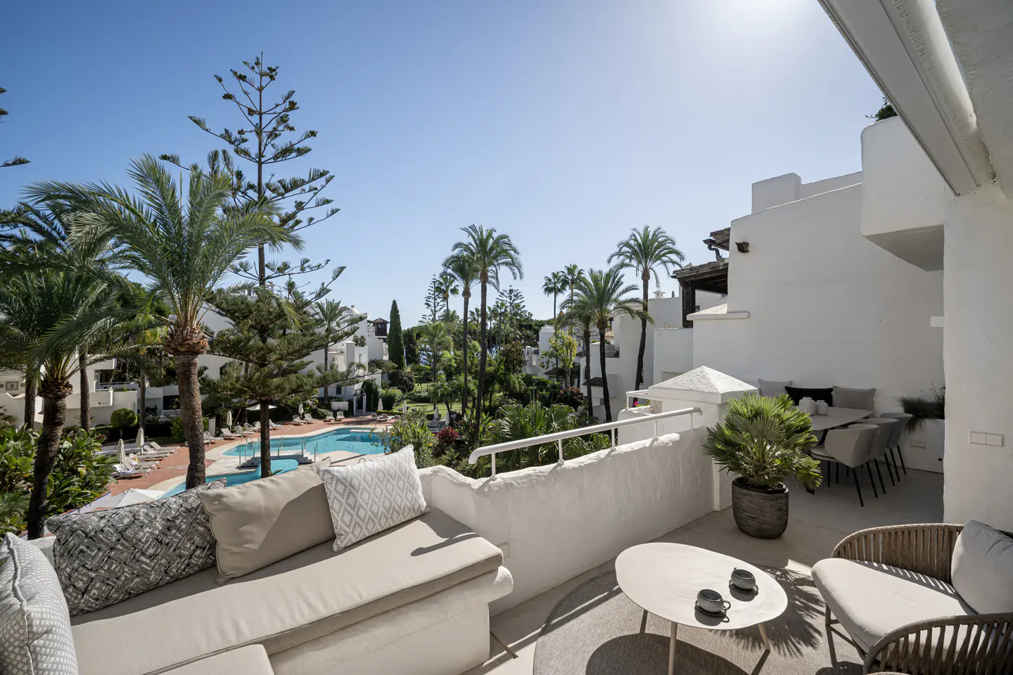 Balcony view of a resort with a pool. White buildings, palm trees, and outdoor furniture are visible.