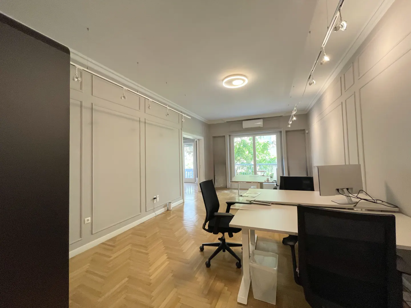 Office space with herringbone wood floors, gray walls, and white desks with black chairs. A large window shows green trees outside.