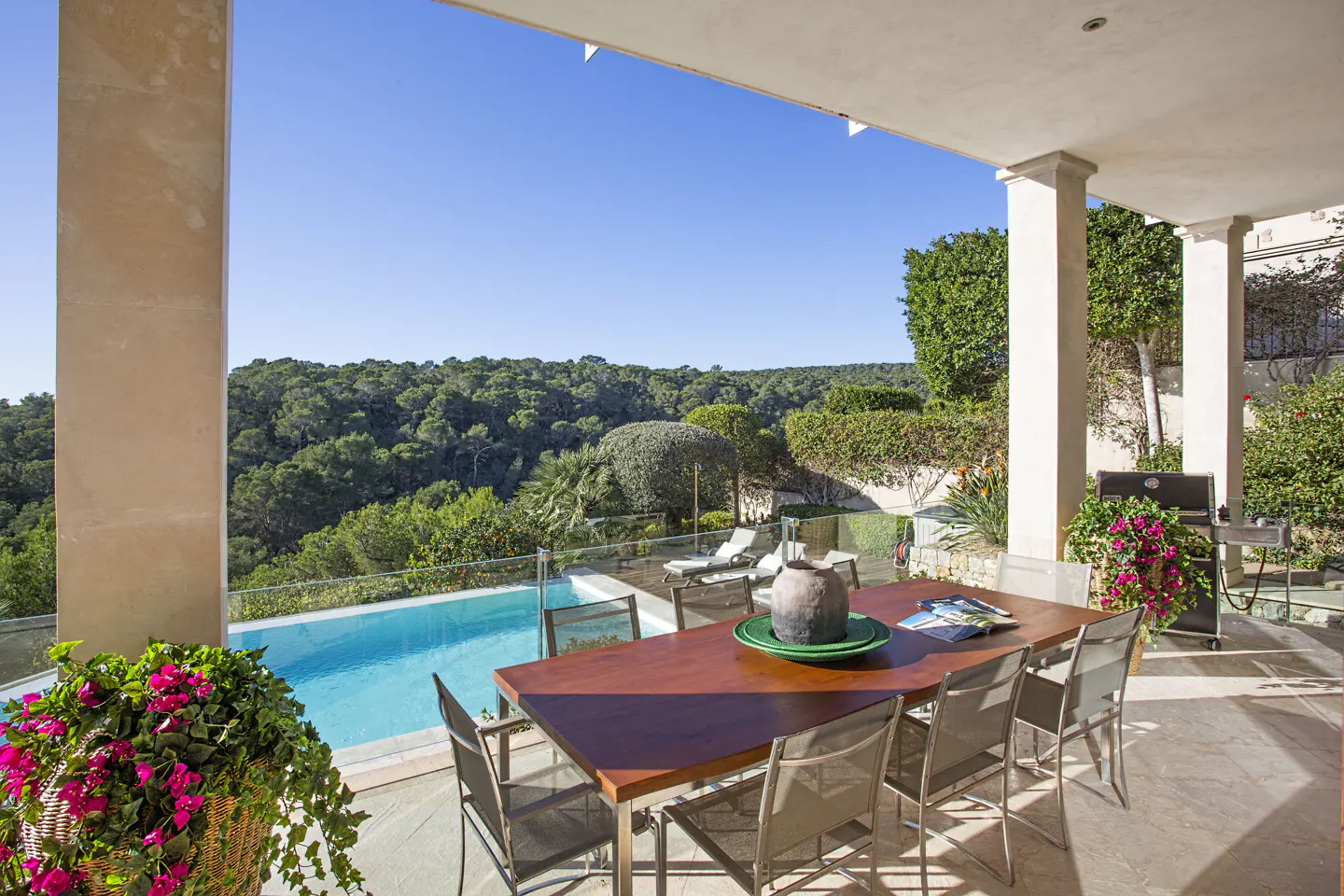 Covered patio with a wood table, metal chairs, pool, and forest view. Grill and pink flowers are on the right.