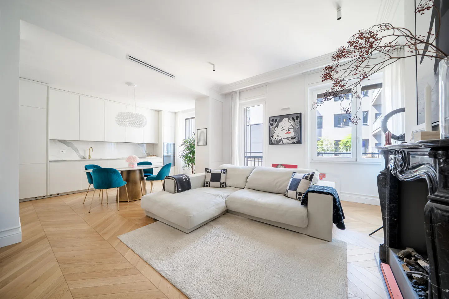 Bright, open-concept living space with white walls, herringbone wood floors, and a large white sectional sofa. A round table with blue chairs sits near a modern white kitchen. A black fireplace is on the right.