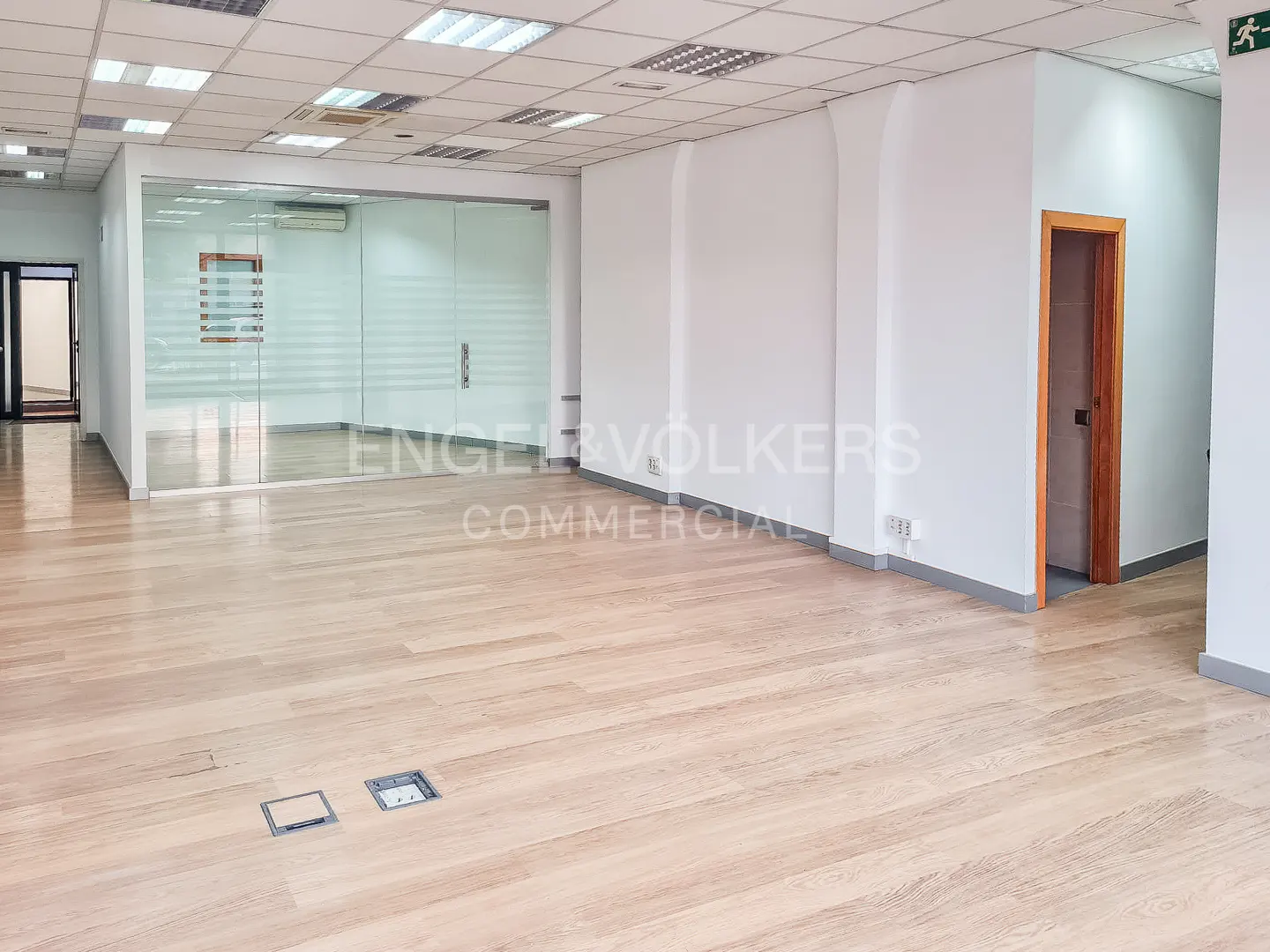 Empty office space with light wood floors, white walls, and a glass-walled room. Recessed lighting in the ceiling.