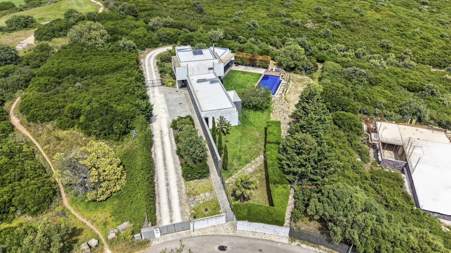 Aerial view of a modern, gray house with a pool, surrounded by lush green trees and a dirt driveway.