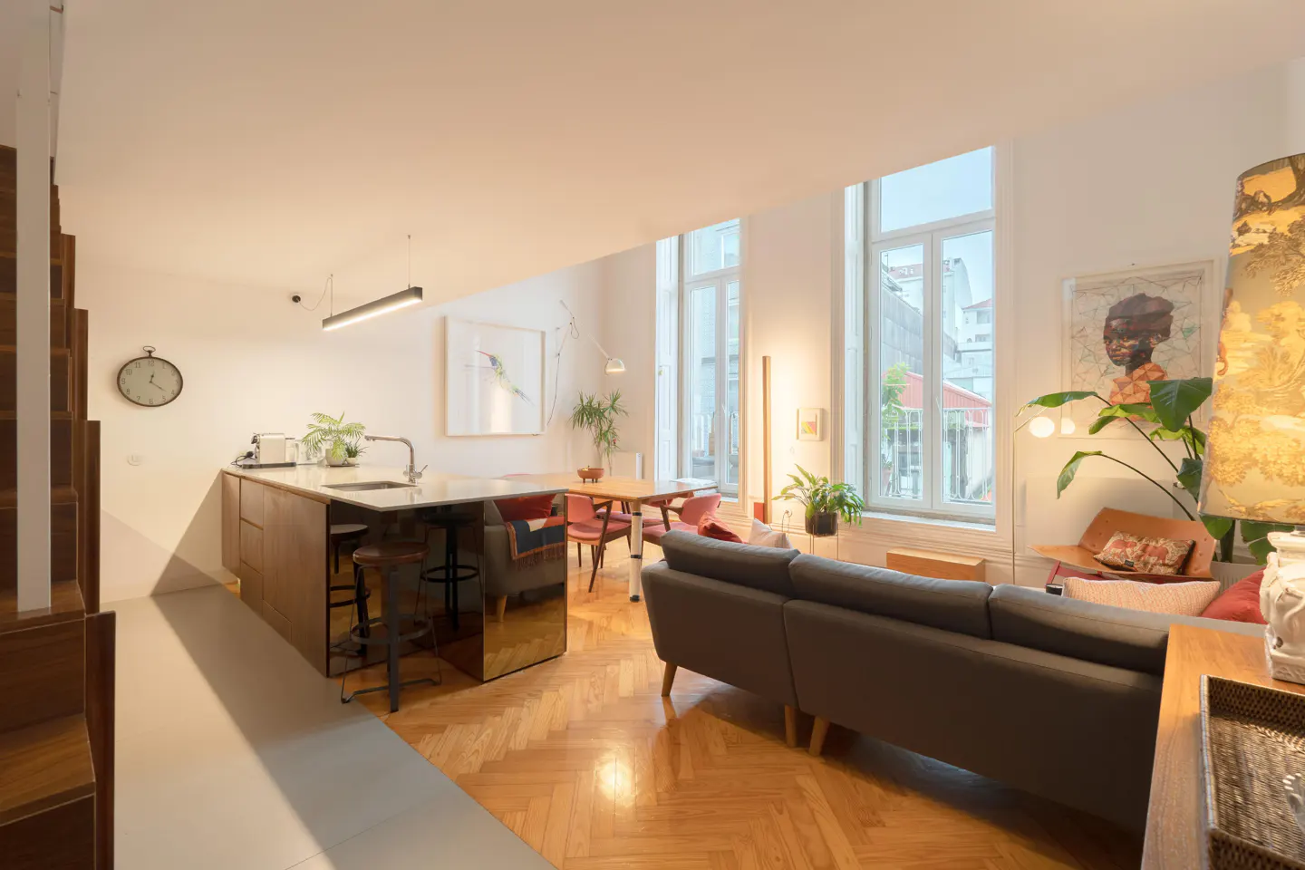 Bright, modern apartment interior with herringbone wood floors. A gray sofa faces large windows. A kitchen island with stools and a dining table are visible. Art adorns the white walls.