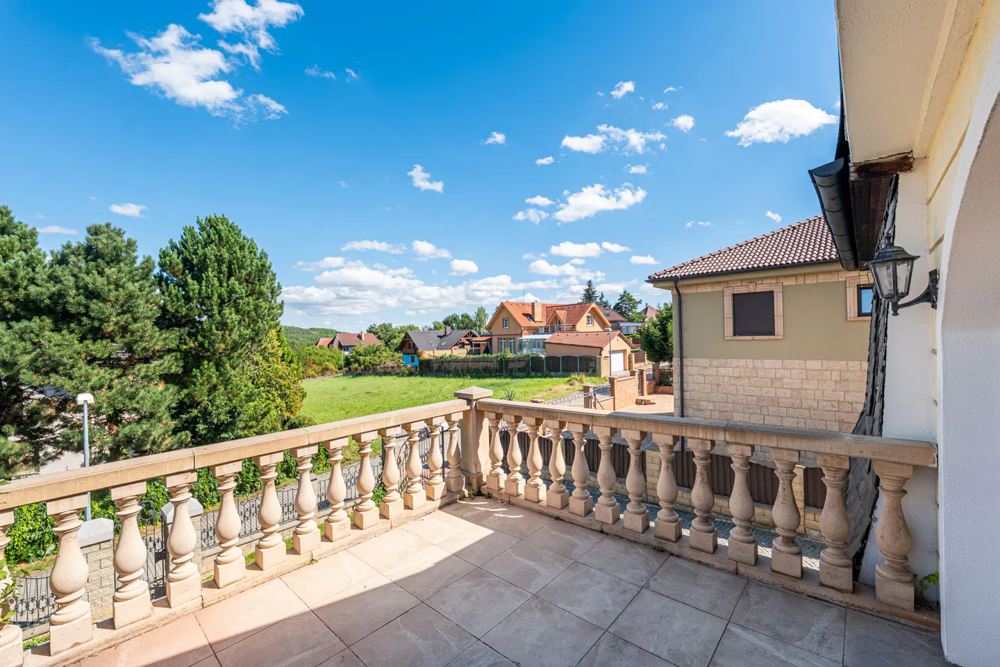 Balcony view with stone balustrade, tile floor, and houses in the background under a blue sky with white clouds.