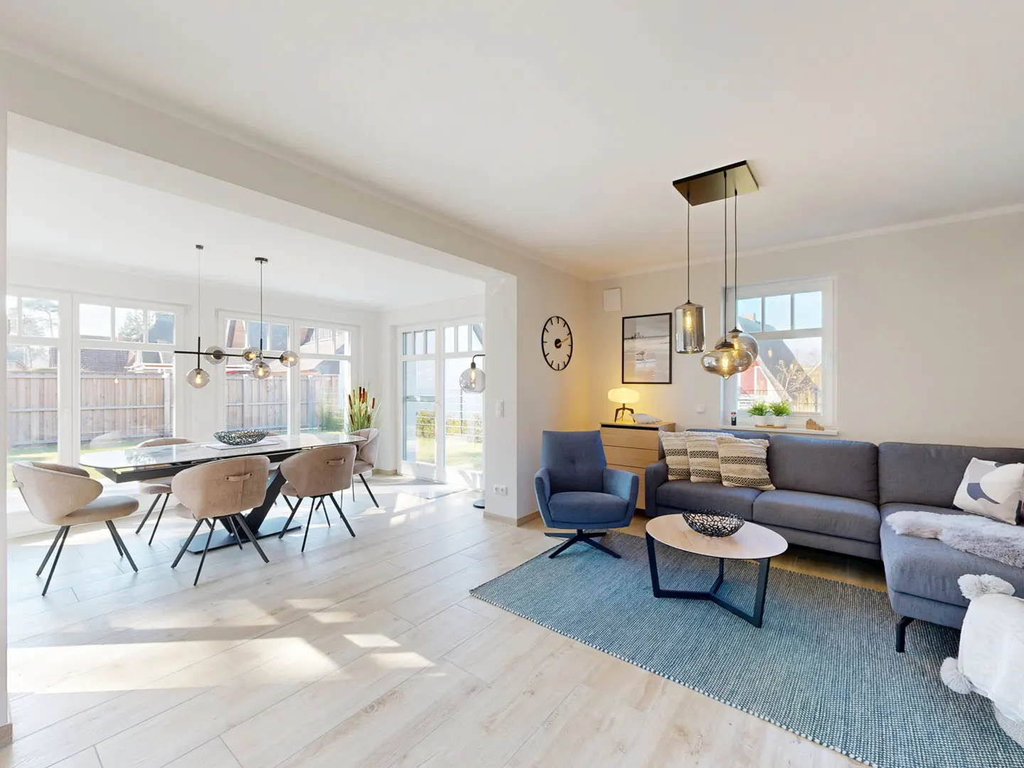 Bright, open-concept living and dining area with a gray sofa, blue chair, and dining table with beige chairs. Light wood floors and large windows.