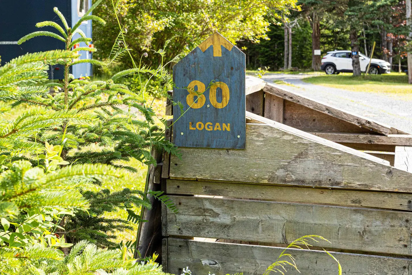 Wooden sign reading "80 Logan" in yellow on a blue background, attached to a weathered wooden structure, with a car parked in the background.