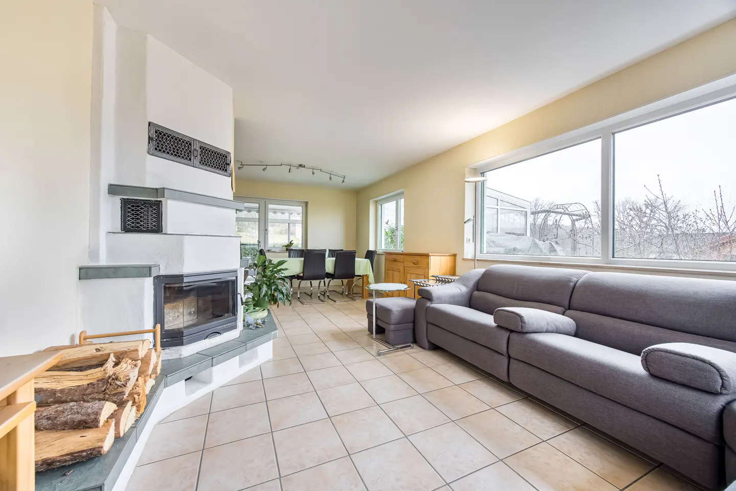 Living room with a gray sofa, fireplace with wood, and a dining table in the background. Tiled floor and large windows.