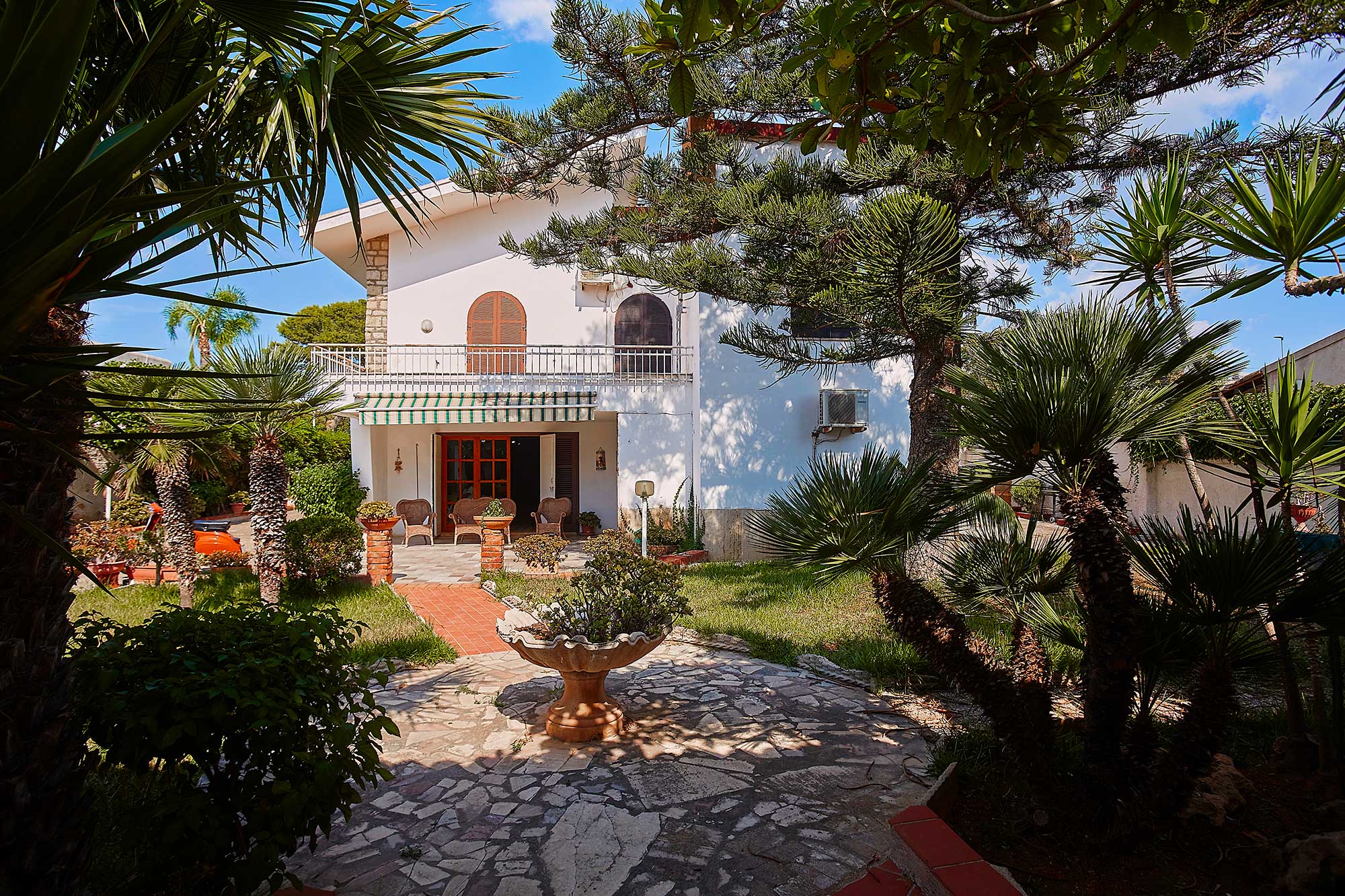 White two-story house with a balcony, patio, and stone fountain, surrounded by lush palm trees and a sunny garden. [AI-generated text]