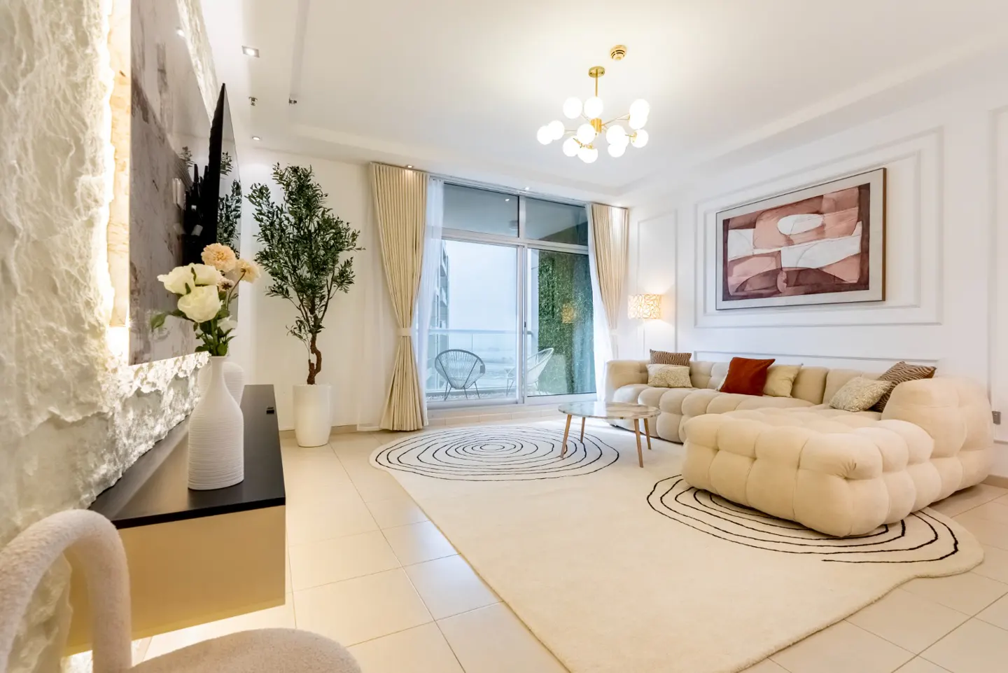 Bright living room with beige sectional sofa, patterned rug, and balcony view. Modern chandelier and artwork add elegance.