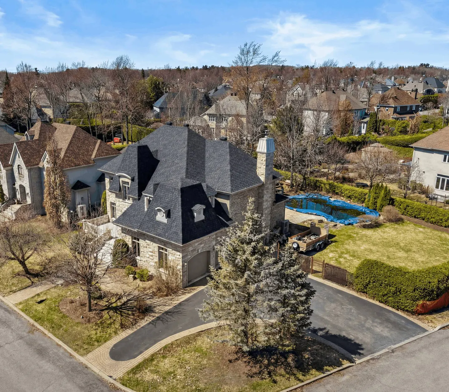 Aerial view of a stone house with a dark gray roof, a pool covered with a blue tarp, and a paved driveway.