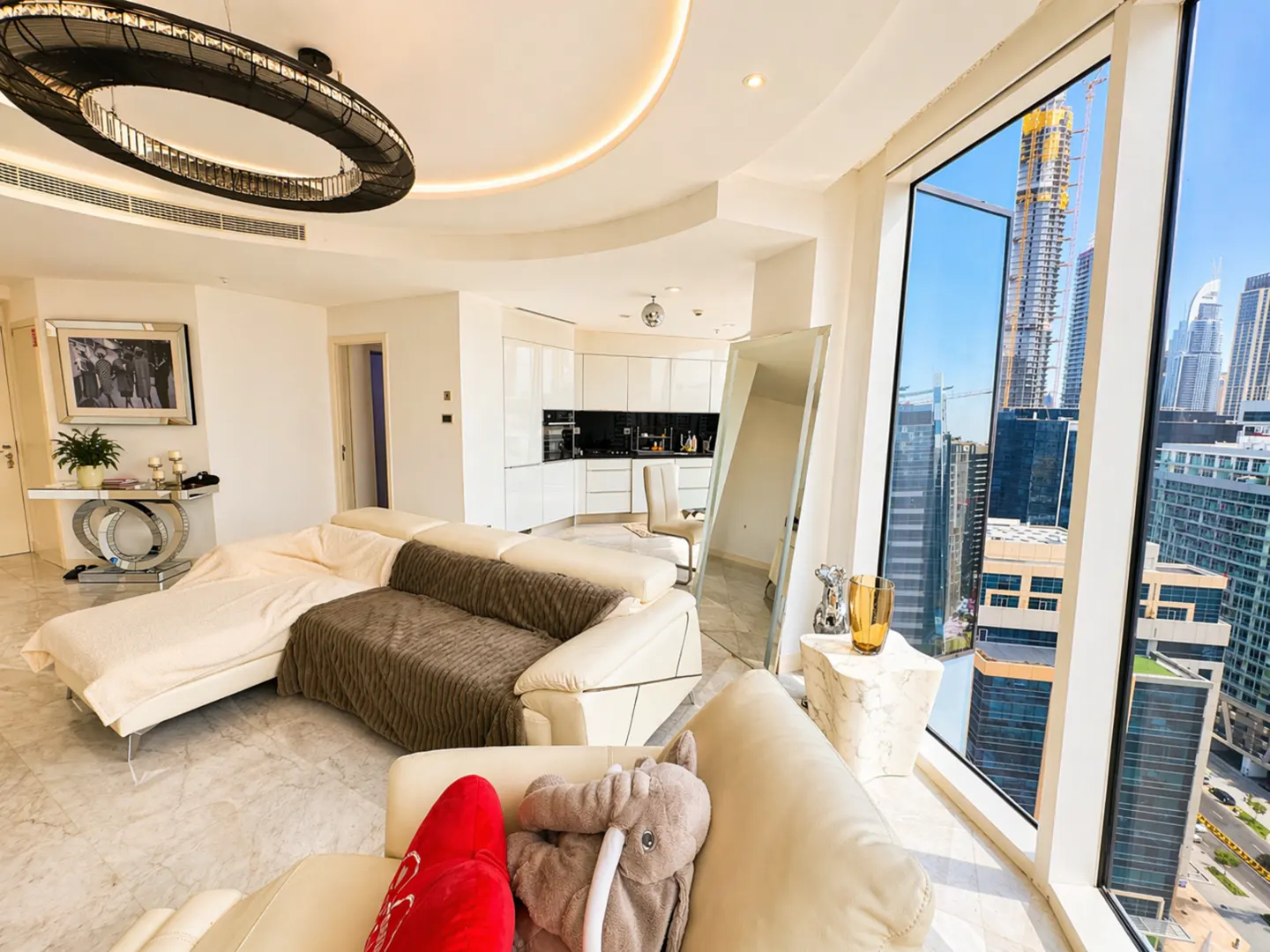 Bright, modern apartment interior with a cream sofa, city view, and open kitchen. A large window shows skyscrapers under a blue sky.